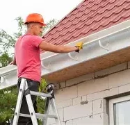 A worker in a hard hat stands on a ladder, repairing a roof gutter.